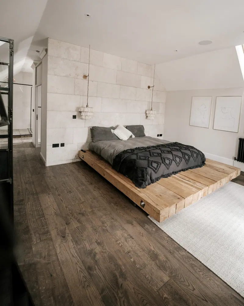 Quarto com cama sobre plataforma de madeira maciça, piso de tábuas escuras e parede de pedra clara.