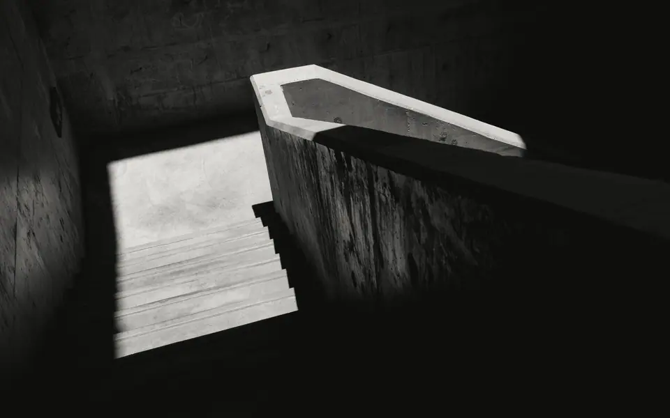 Escada de concreto em penumbra com um recorte de luz forte sobre os degraus, em preto e branco. Imagem de referência.