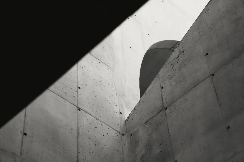 Paredes de concreto aparente atravessadas por uma sombra diagonal, com um volume curvo ao fundo, em preto e branco. Imagem de referência de banco de imagens.