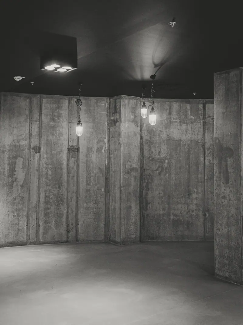 Sala de paredes de concreto com marcas de fôrma e luminárias pendentes, em preto e branco.