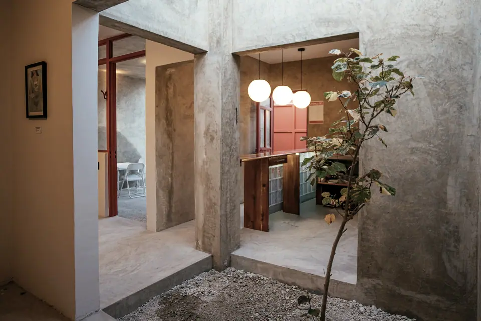 Pátio interno com paredes de concreto, piso de pedriscos e uma pequena árvore, com ambientes iluminados ao fundo.