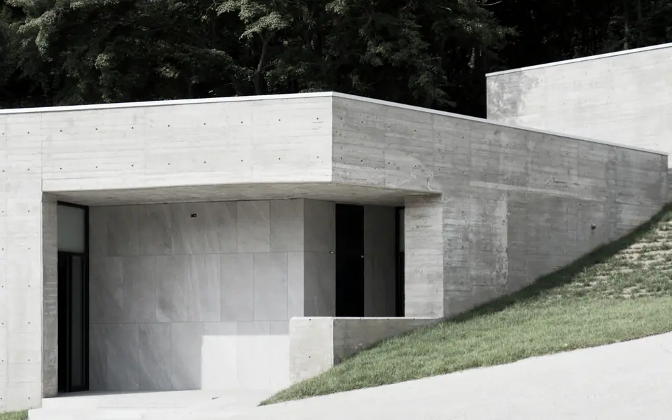Volume baixo de concreto aparente encaixado em um talude gramado, com árvores altas ao fundo.